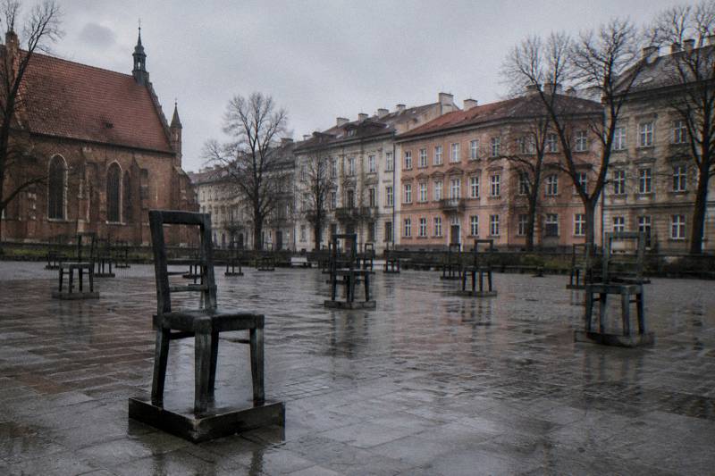 Mañana nublada de principios de primavera en la Plaza de los Héroes del Gueto, en Podgórze, Cracovia, Polonia: filas de grandes sillas de bronce vacías, espaciadas sobre el pavimento de la plaza abierta, piedra mojada, bloques de viviendas sencillos y árboles sin hojas al fondo, una visión documental del lugar, sin personas en el encuadre.