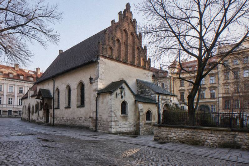 Bright cold midday in late autumn outside the Old Synagogue in Kazimierz, Krakow, Poland, a low pale Gothic-Renaissance building with a tall steep roof and a stepped decorative parapet, bare tree branches across a pale sky, a patch of damp cobbles in the foreground, nobody in frame.