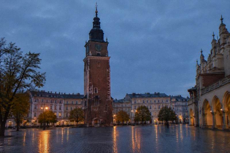 Blue hour on an autumn evening in the Main Market Square in Krakow, Poland, the lone tall brick Town Hall Tower standing by itself on the cobbles with a slight lean, damp paving reflecting the first warm streetlights, the edge of the Cloth Hall just visible at the side, two tiny distant figures.