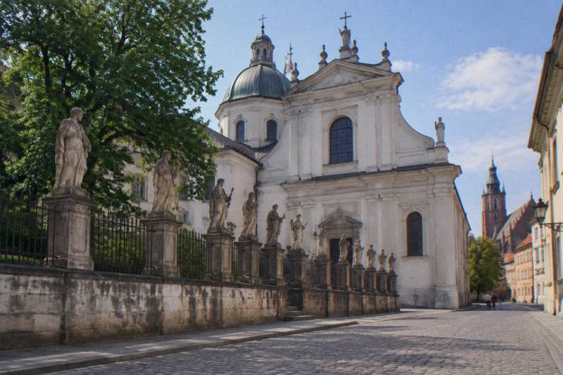 Tarde soleada de principios de verano frente a la gran iglesia barroca blanca de San Pedro y San Pablo, en Cracovia, Polonia: un muro bajo de piedra rematado por una hilera de figuras de piedra a tamaño real, con la cúpula y la fachada lisa de la iglesia al fondo, un tramo de la calle empedrada Grodzka en primer plano, dos figuras diminutas a lo lejos.