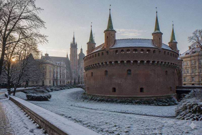 Mañana fría y despejada tras una nevada reciente en la Barbacana de Cracovia, Polonia: la bastión circular fortificado de ladrillo rojo con sus esbeltas torres y troneras visto desde el foso herboso, una fina capa de nieve en el césped y los árboles, sol bajo y pálido, sin nadie en el encuadre.