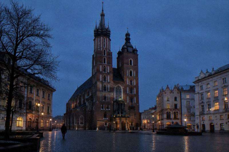 Winter dusk at St Mary's Basilica in Krakow, Poland, seen from the small square at its foot, the two uneven brick Gothic towers rising dark against a deep blue sky, a few lit windows in the neighbouring tenements, cold damp cobbles, one tiny distant figure with their back to the camera.