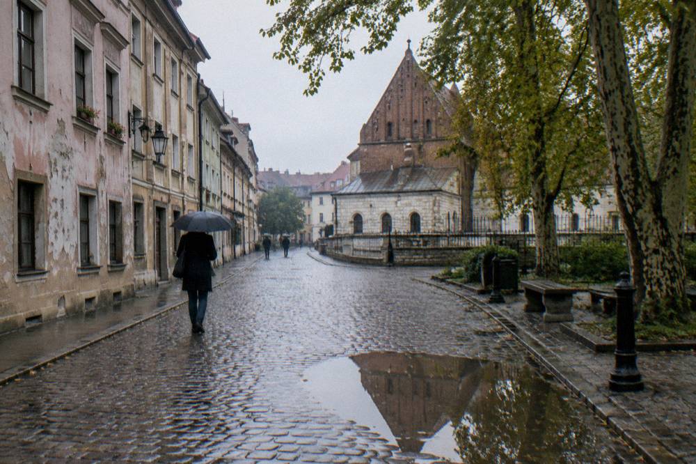 Rainy cobblestone street with worn pastel townhouses, plane trees, a brick synagogue gable, puddles and a person under an umbrella.