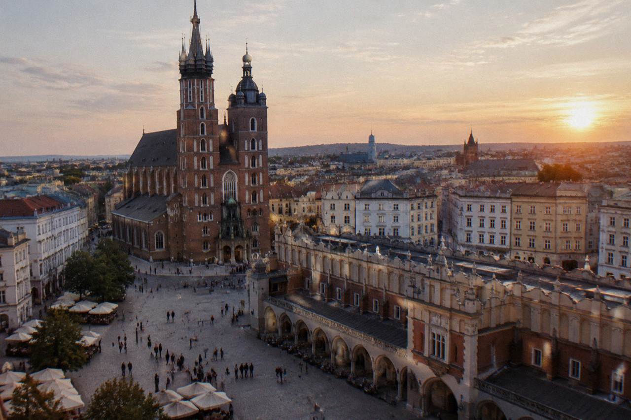 Golden hour view over Krakow Main Market Square with St Mary's Basilica, the Cloth Hall, rooftops, cobbles, and distant pedestrians.