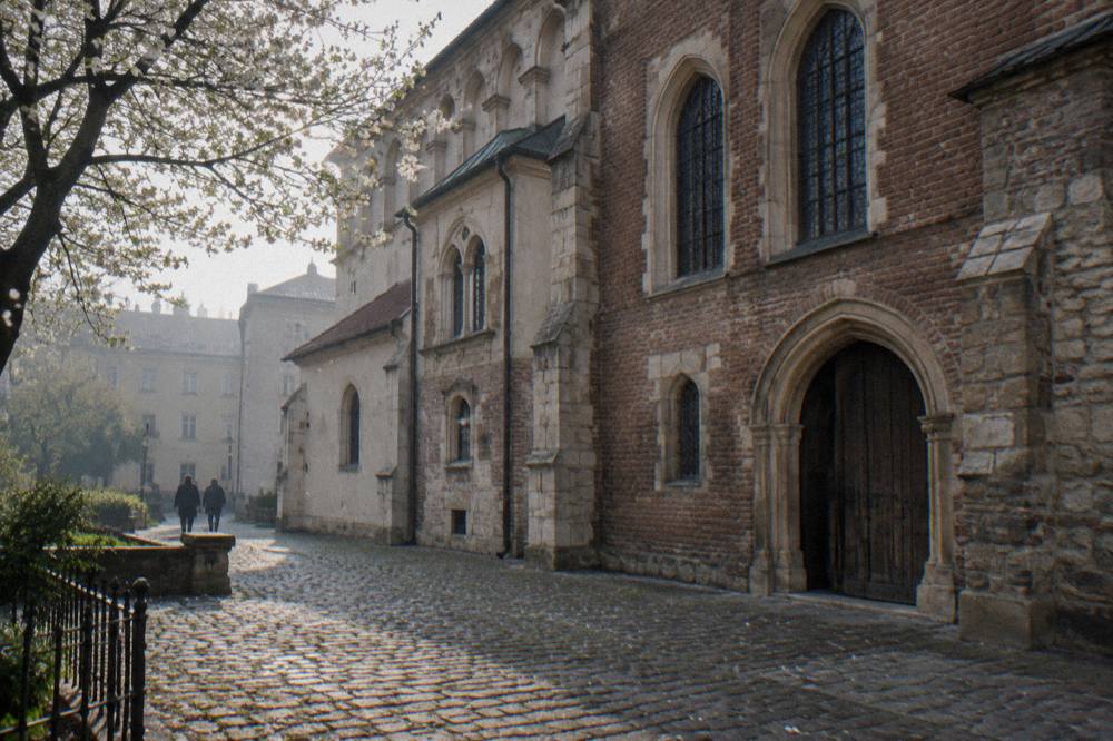 Cobbled courtyard beside a brick Gothic building with arched windows, a dark wooden door, blossom branches, and two distant walkers.