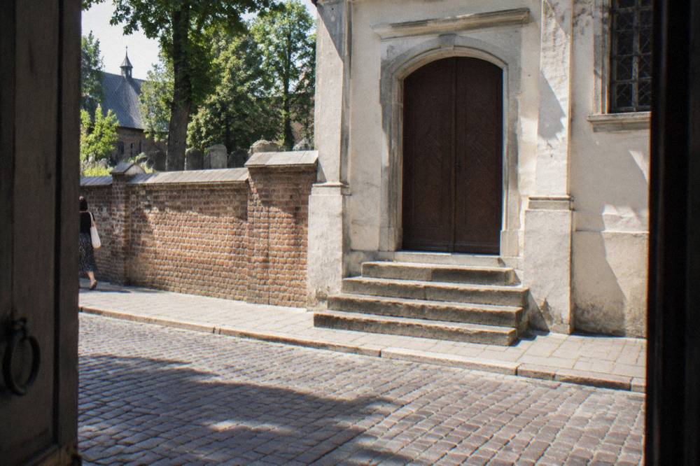 Closed wooden synagogue door above worn stone steps beside a low brick wall on a cobbled street in Kazimierz.