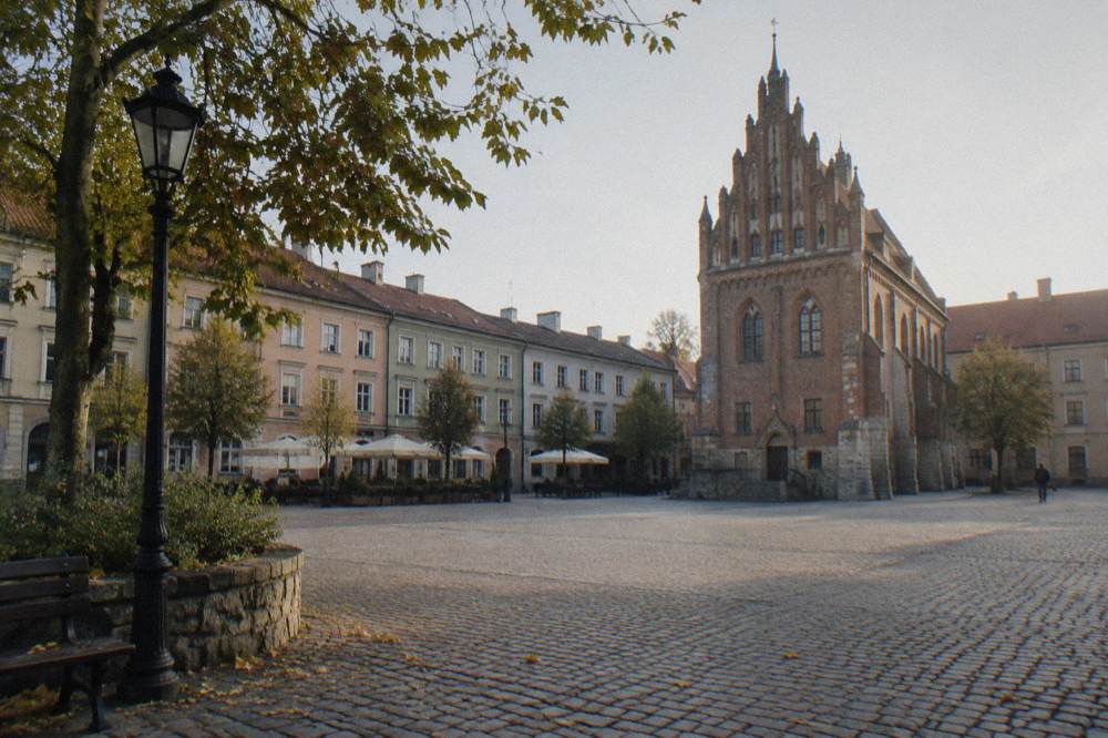 Cobbled square with pastel townhouses, a Gothic brick town hall, trees, benches, and a few distant pedestrians.
