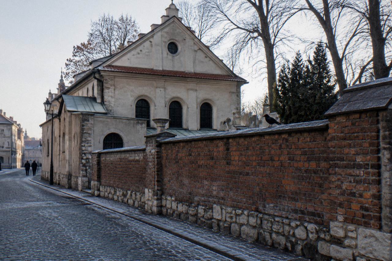 Old synagogue facade and brick cemetery wall line a frosty cobbled street with bare trees and a crow on the wall.