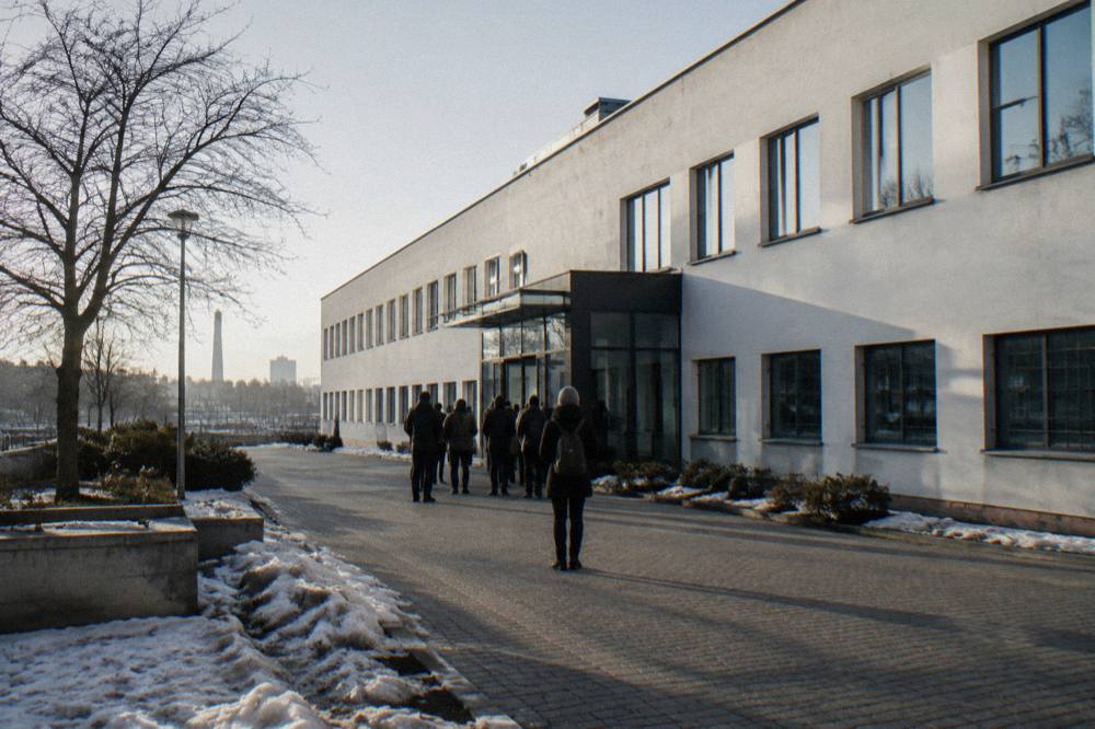 A snowy paved forecourt outside a long white factory building with a small line of visitors near the entrance.