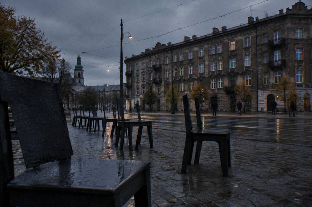 Large empty steel chairs stand on wet cobblestones beside tram rails and old tenement facades at dusk.