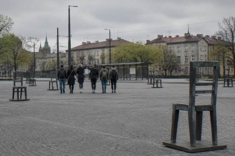 Five people walk across Ghetto Heroes Square among large steel chair sculptures, with a tram shelter and old tenements beyond.