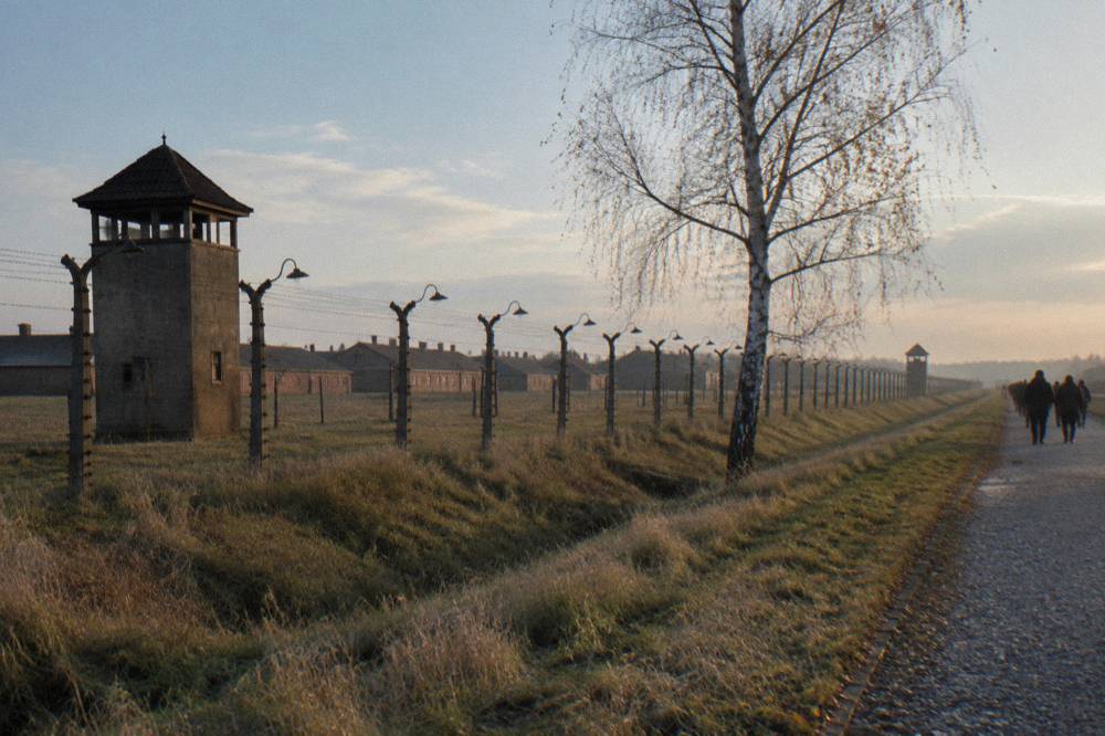 A concrete watchtower and barbed wire fence line run across dry grass beside a birch tree at Birkenau.