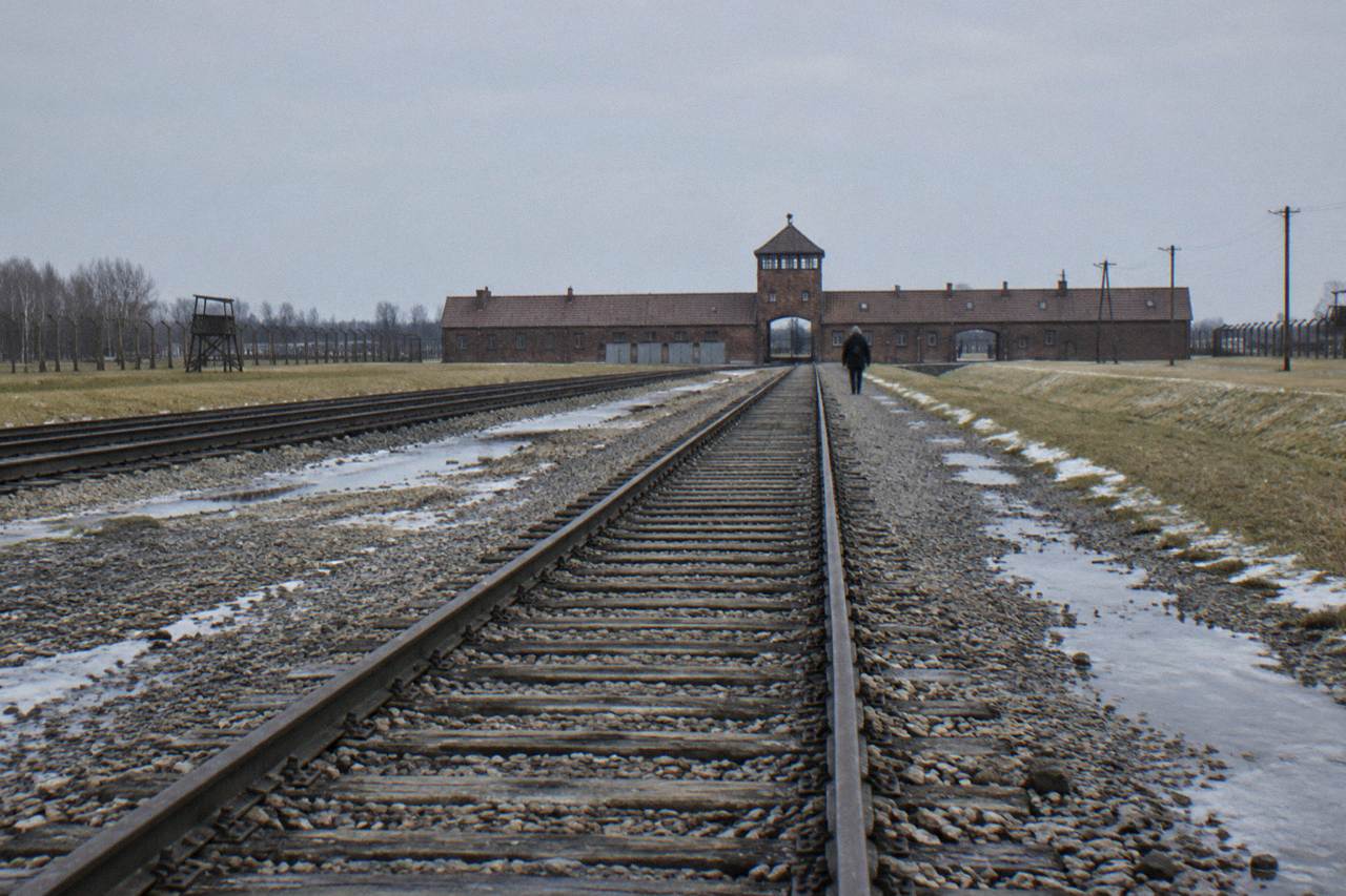 Rail tracks lead across gravel and snow patches toward the brick Birkenau gatehouse under a pale sky, with one distant visitor.