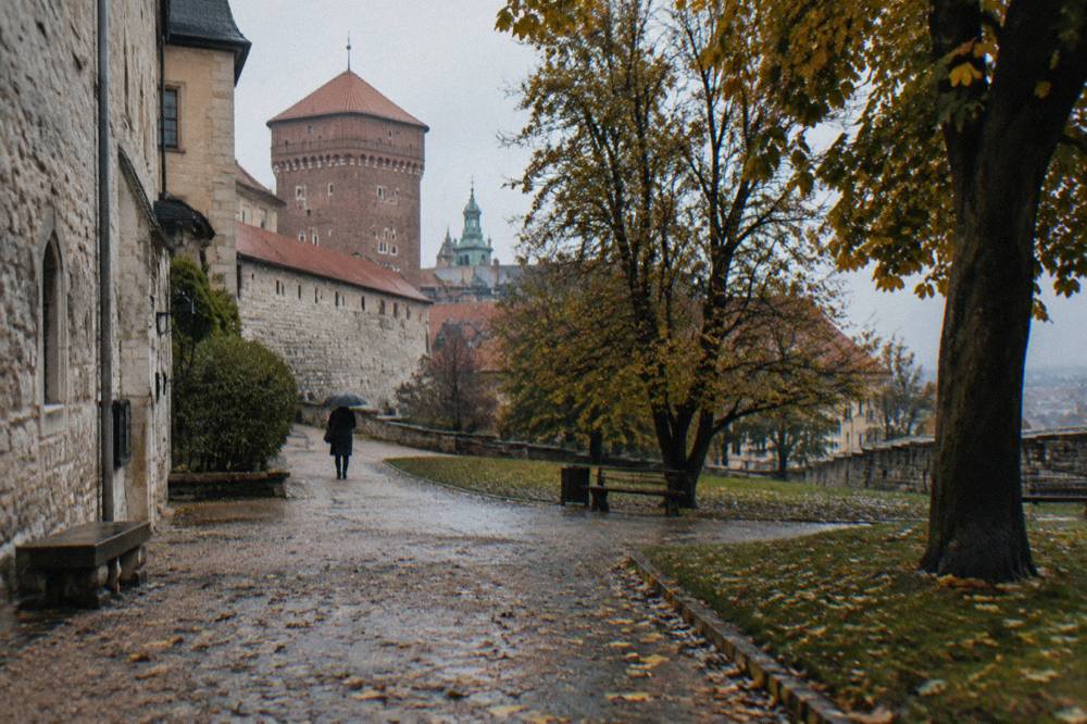 Wet gravel path on Wawel Hill passes stone castle walls, autumn trees and a distant person with an umbrella.