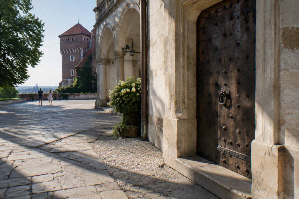 Closed studded wooden door in a stone castle wall beside arcades, with flagstones, gravel, trees and a brick tower in the background.