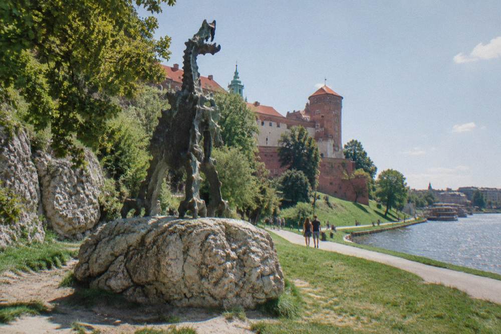 Bronze Wawel Dragon statue on a stone plinth beside the Vistula, with Wawel Castle and riverside path behind it.
