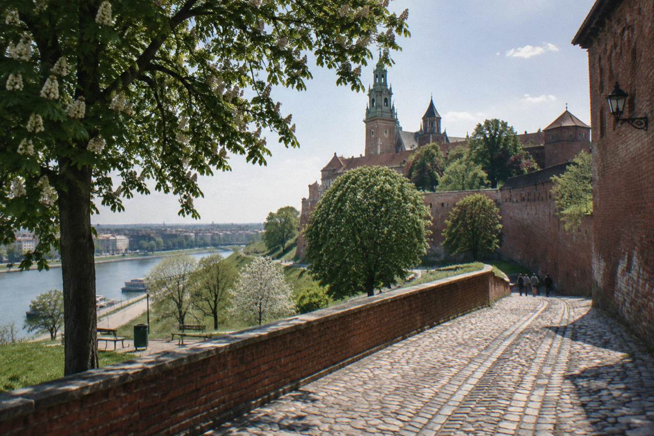 Cobbled path beside red brick Wawel Castle walls overlooks the Vistula under leafy chestnut branches.