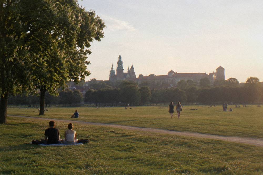 People sit and walk on a broad grassy meadow with trees, a dirt path, and distant Krakow church and castle towers at sunset.