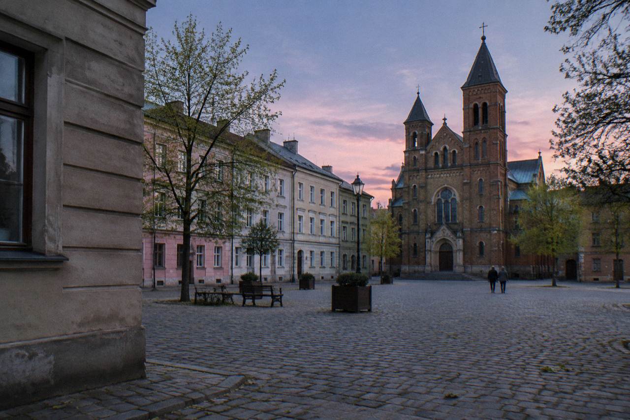 Empty cobblestone square with pastel tenements, benches, trees and a brick twin-towered church under a pink evening sky.
