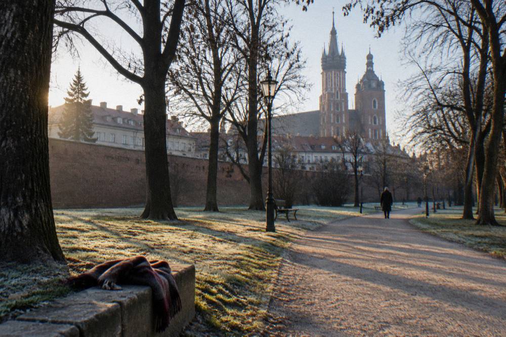 Frosty Planty park path with bare trees, a scarf on a stone kerb, and Krakow church towers rising beyond a brick wall.