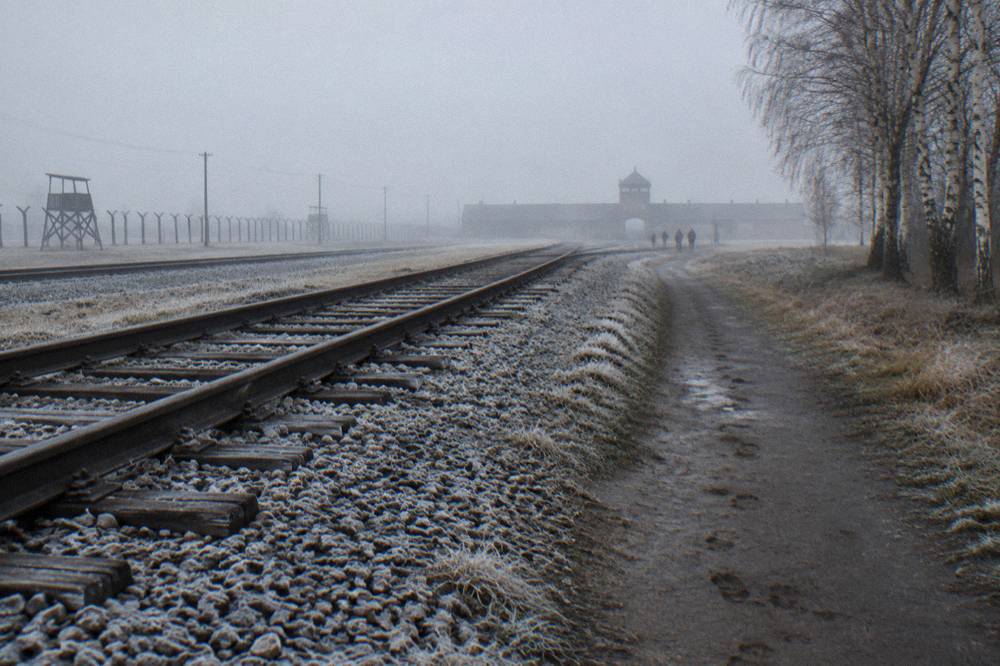 Vías de tren heladas y un camino embarrado llevan hacia la puerta de Birkenau entre la niebla, con vallas, una torre de vigilancia y árboles desnudos.