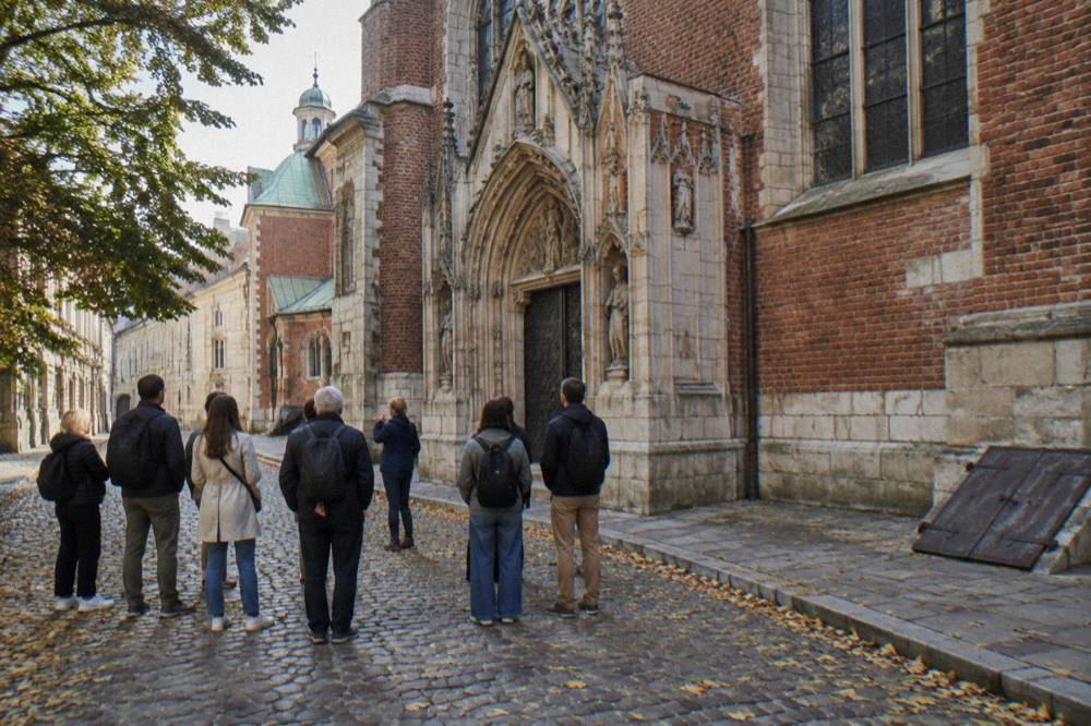 A small walking tour group stands on a cobbled street beside a Gothic brick church facade with trees and fallen leaves.