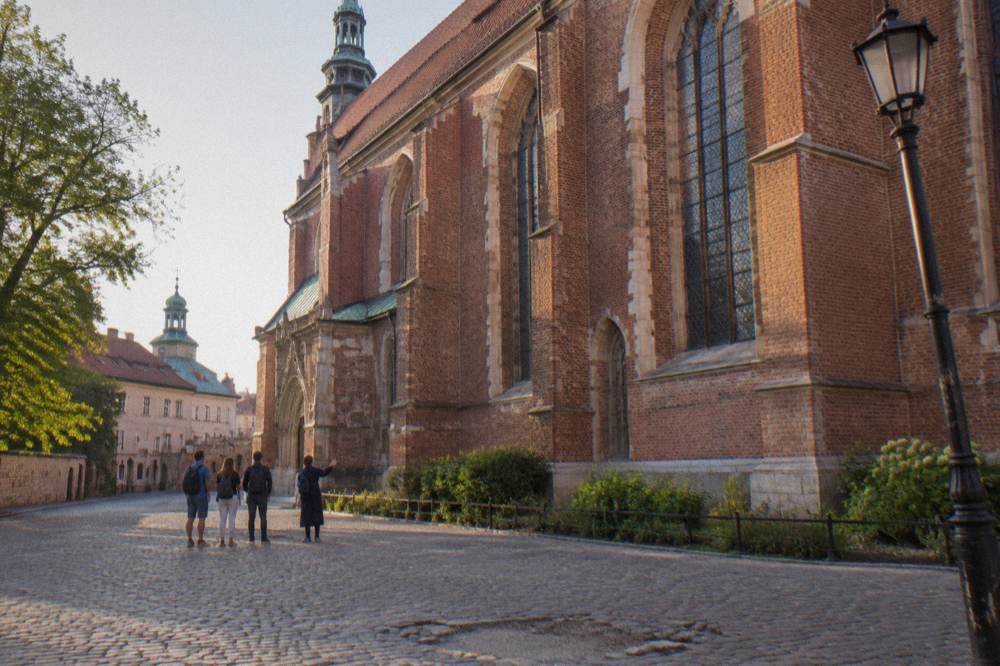 A guide gestures to three visitors on cobbles beside a tall red brick Gothic church facade and a leaning lamppost.