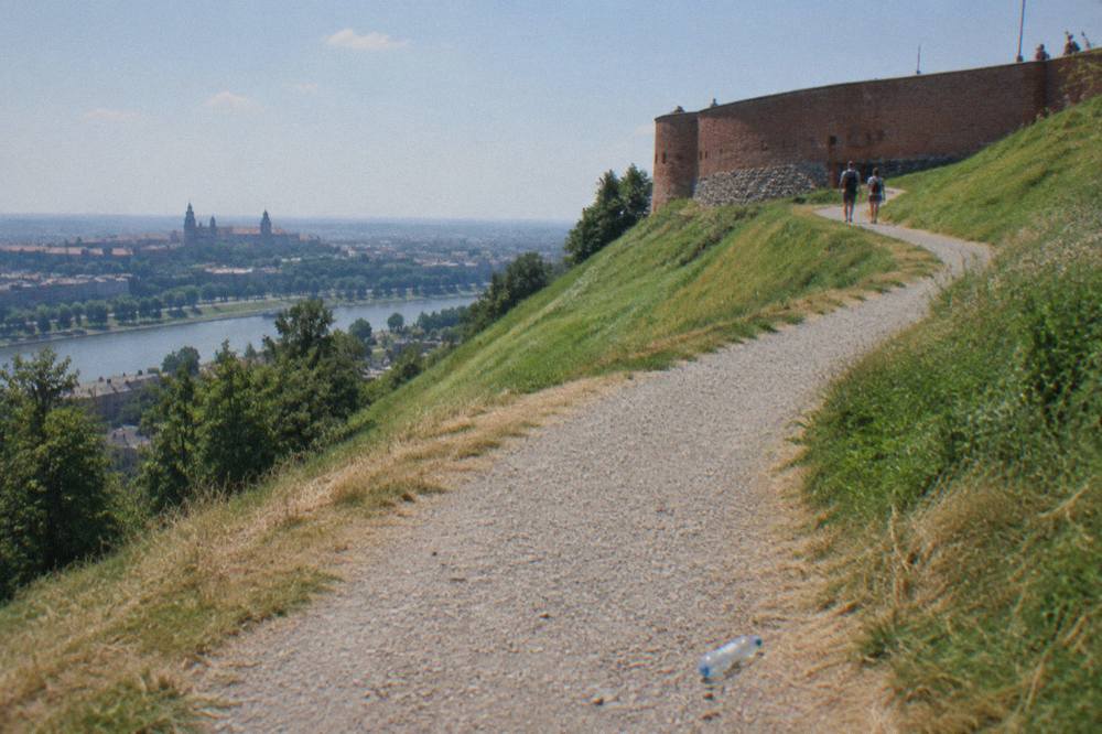 A gravel path curves up a grassy mound toward a brick fort wall, with distant walkers, a river valley, and a plastic bottle on the path.