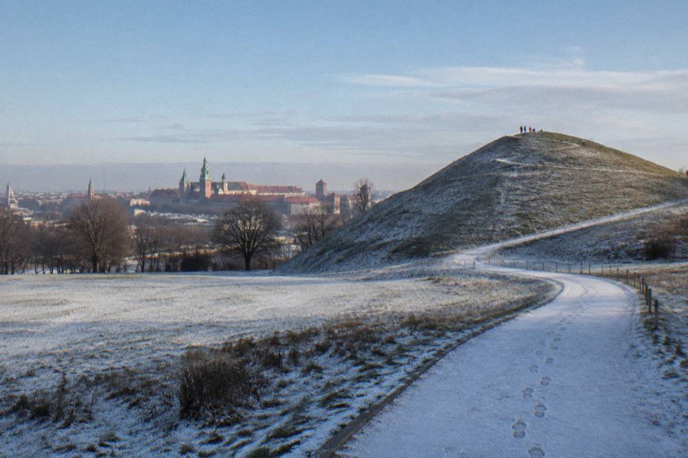 Snow-dusted Krakus Mound rises beside a curving path with footprints, with distant Krakow rooftops and spires beyond the parkland.