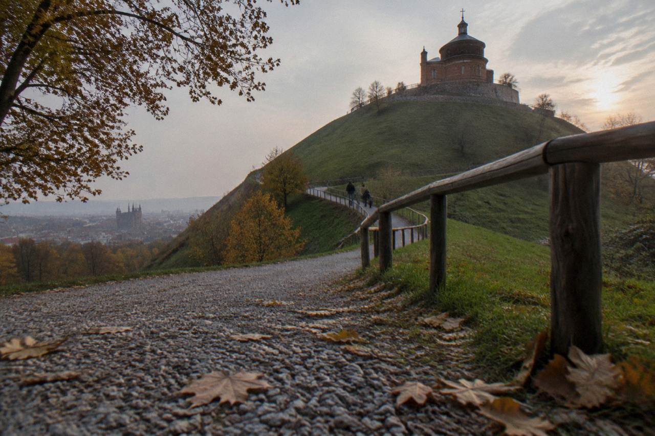 A gravel path with fallen leaves spirals up a grassy mound to a brick chapel, with a wooden handrail and hazy city view.