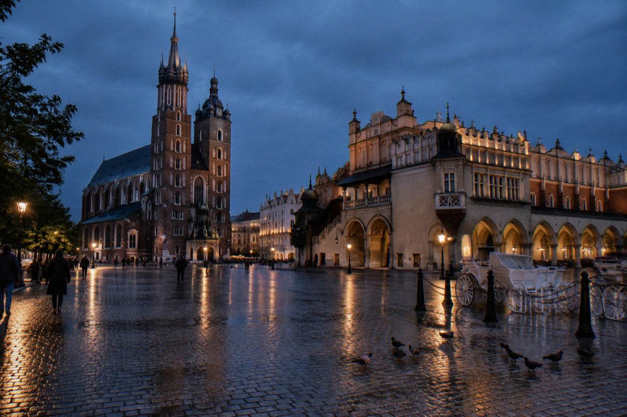 Wet cobbles reflect lamps in Krakow Main Market Square, with St Mary's Basilica, the Cloth Hall, pigeons and a white carriage at dusk.