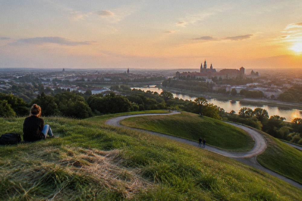 Grassy hilltop with a winding gravel path, seated visitors, the Vistula, and Krakow skyline at sunset.