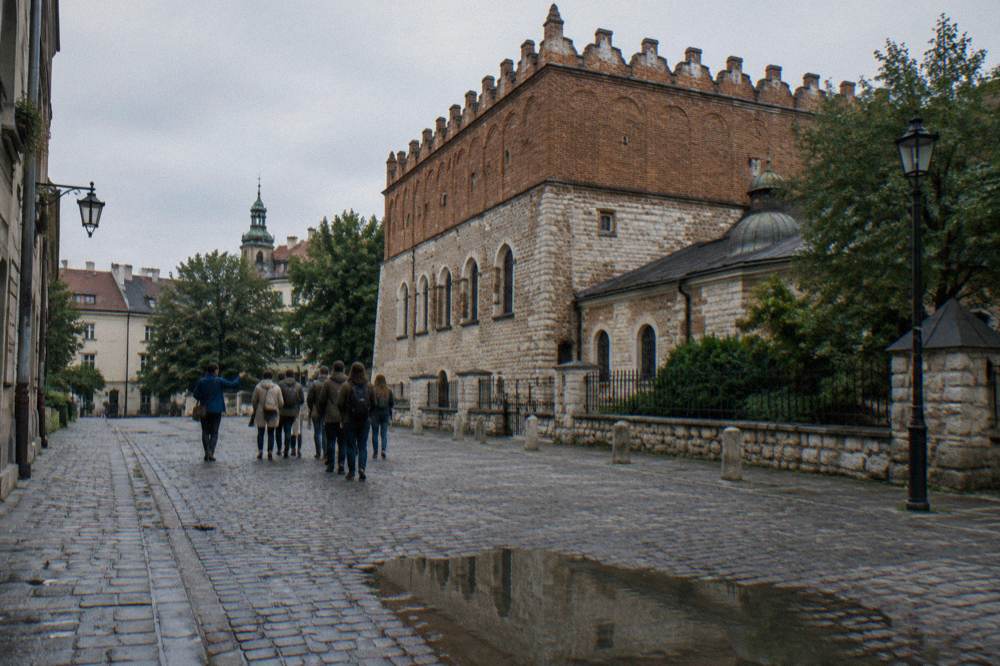 A small group walks away on wet cobbles beside a crenellated brick and stone synagogue with trees and a puddle.