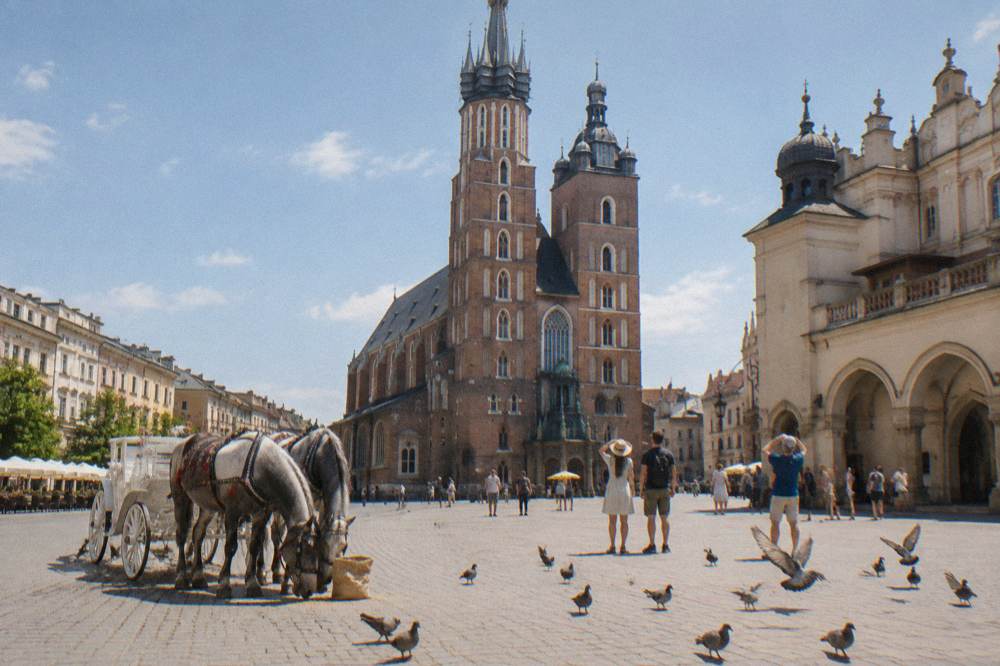 People stand on Krakow Main Market Square near a horse carriage, pigeons, St Mary's Basilica and the Cloth Hall.