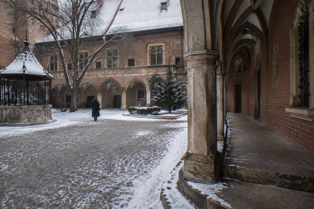 Snowy cobbled courtyard with brick Gothic arcades, a small pavilion, bare trees, and one distant person walking away.