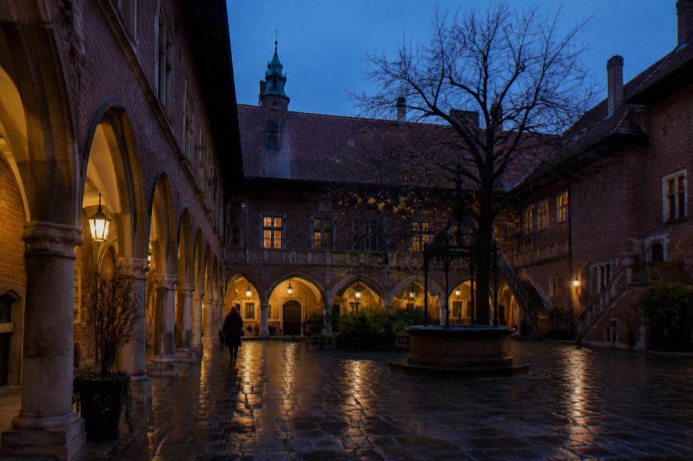 Lamp-lit brick arcades surround a wet cobbled university courtyard at blue hour with one distant figure crossing.