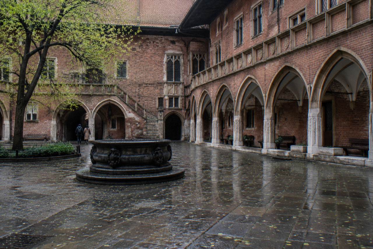 Wet Gothic brick university courtyard with pointed arcades, a stone well, benches, a tree, and two distant people.