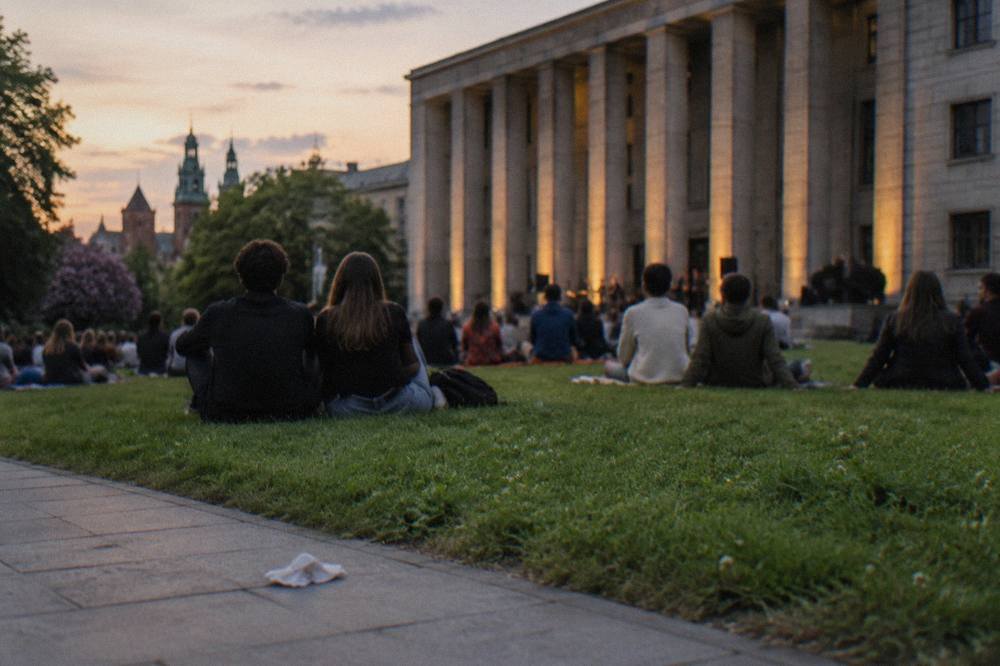 People sit on grass at dusk outside a lit colonnaded museum facade, with a crumpled napkin on the pavement.