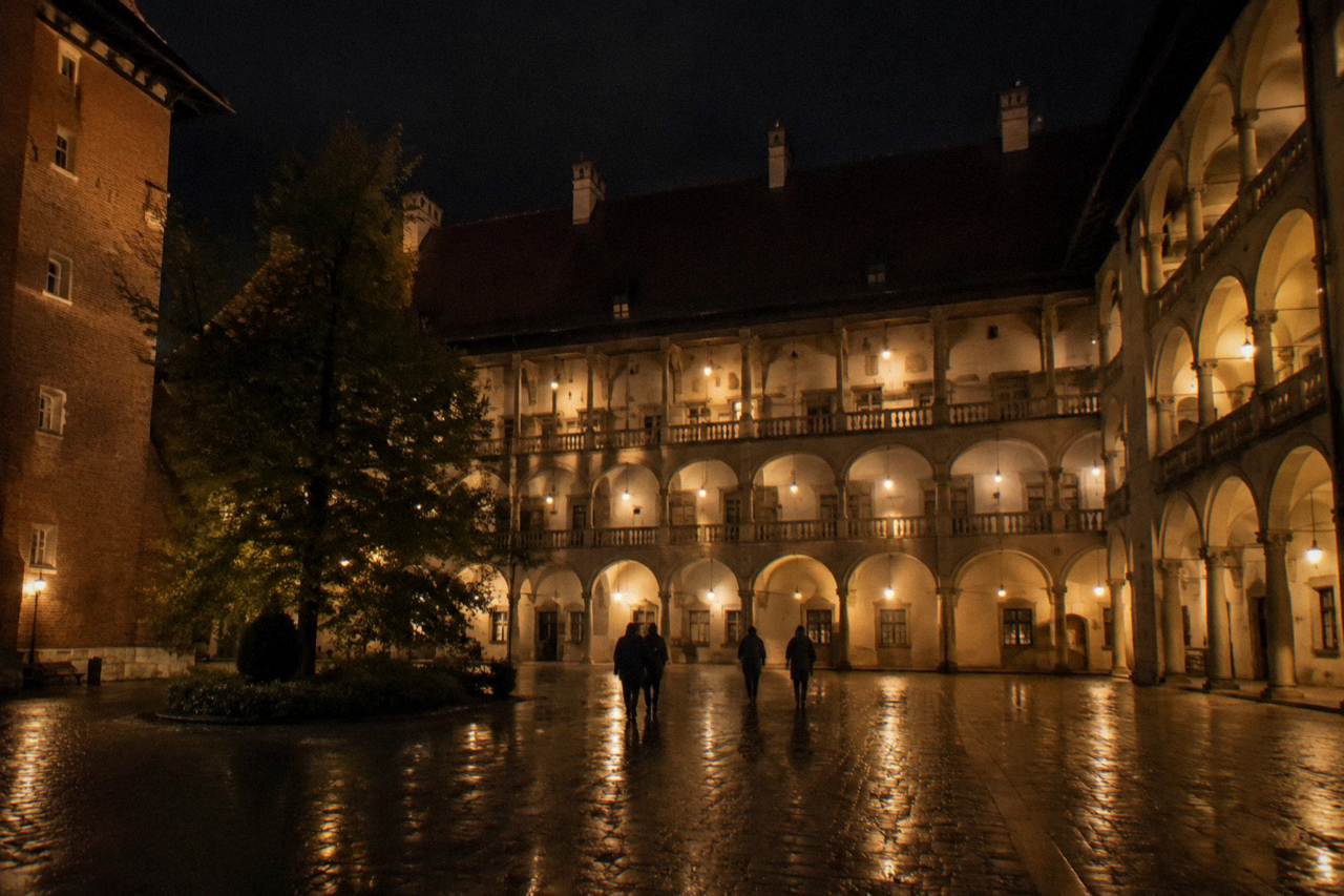 Warm lights illuminate Wawel Castle arcades at night as visitors cross a wet stone courtyard.