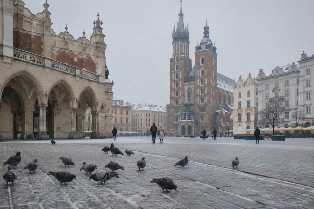 Pigeons peck on snow-dusted cobbles as bundled visitors cross Krakow Main Market Square near the Cloth Hall and St Mary's Basilica.