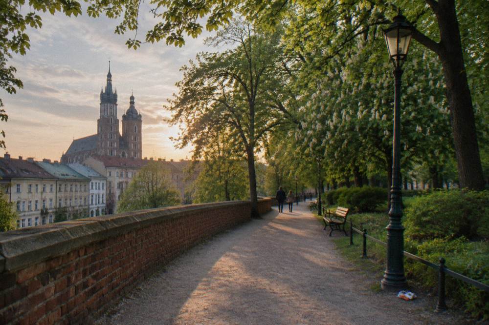 Tree-lined gravel promenade beside a brick wall, with benches, lampposts, distant walkers and Krakow church towers beyond old facades.