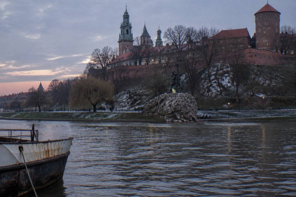 Wawel Hill rises above the Vistula at winter dusk, with a dark dragon statue near the rocky bank and a rusted boat hull in front.