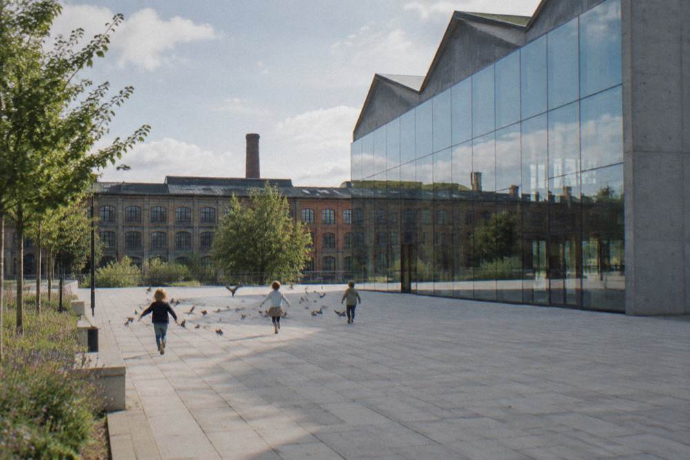 Children chase pigeons across a paved museum square beside a glass facade, with old brick factory buildings behind.