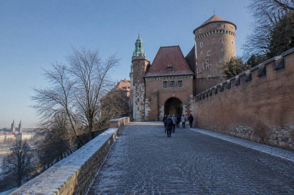 Visitors walk up a frosty cobbled ramp toward a brick castle gate and round tower on Wawel Hill under a clear blue sky.