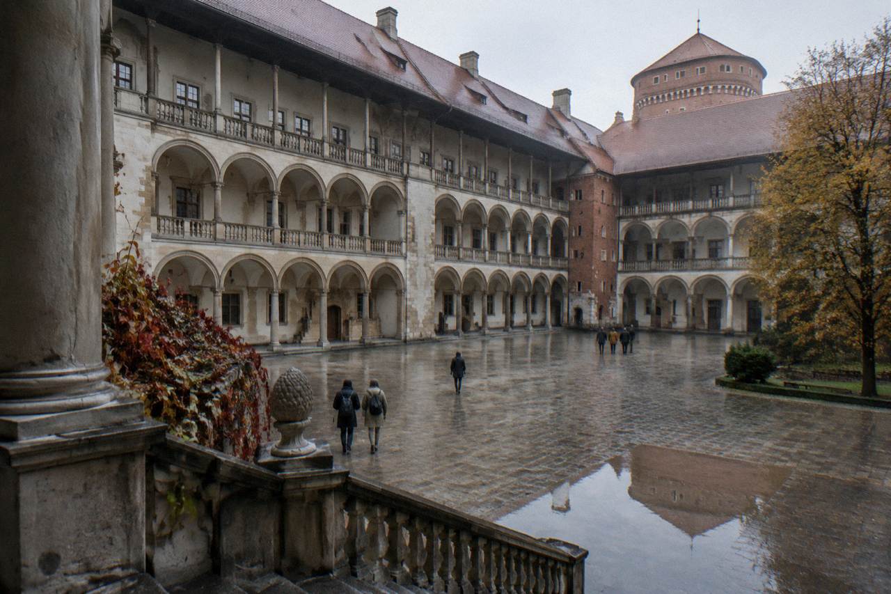 Rain-wet Wawel Castle courtyard with Renaissance arcades, a stone stair, autumn trees, and a few distant people.