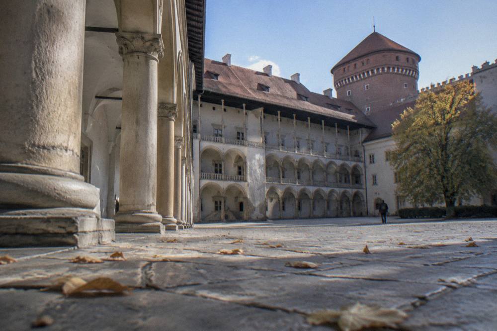 Low view across Wawel Castle courtyard with stone columns, arched galleries, scattered leaves, and a round brick tower.