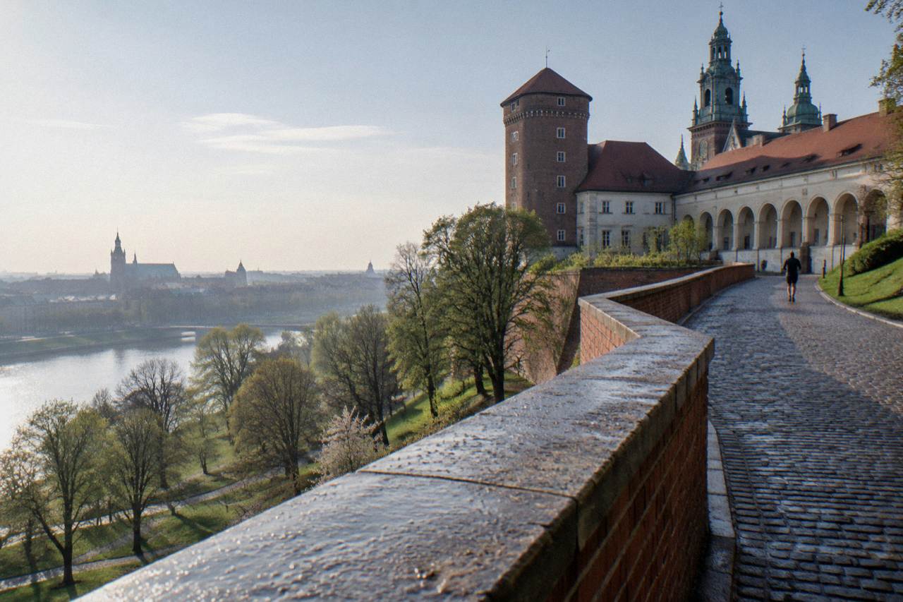 Cobbled path beside a dew-covered brick wall on Wawel Hill, with castle arcades, towers, trees, and the Vistula below.
