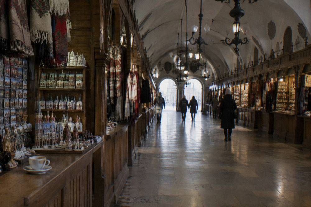 Sala de comercio abovedada con puestos de souvenirs de madera a ambos lados, un pasillo de piedra pulida, lámparas colgantes, visitantes a lo lejos y una taza sobre el mostrador.