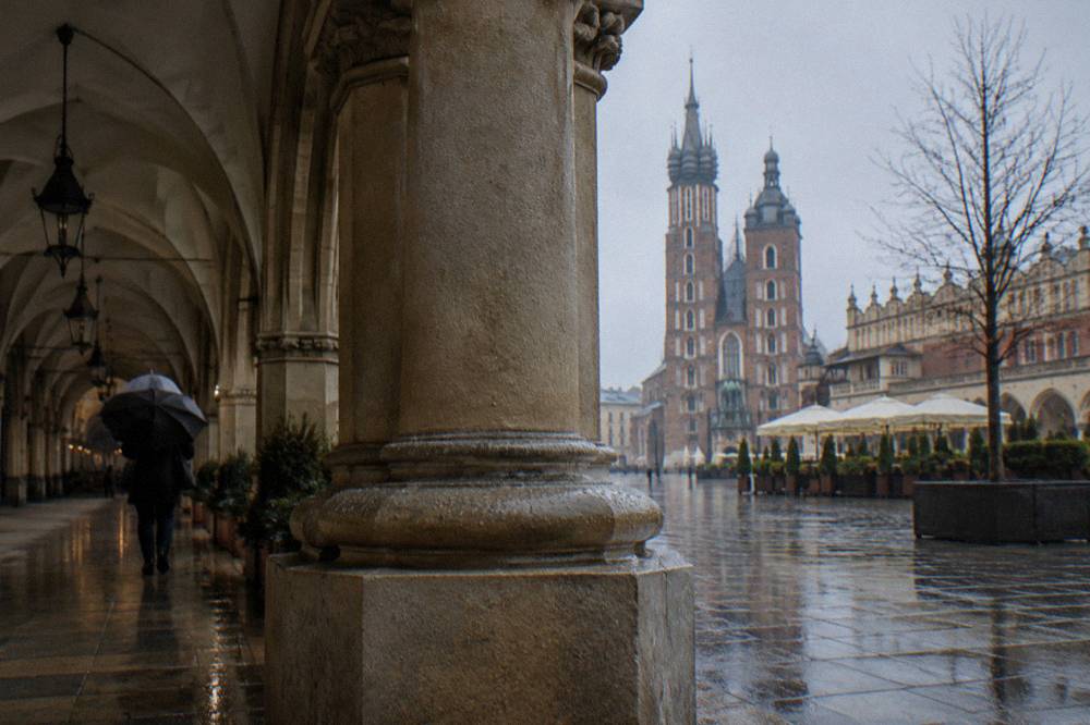 Columna de la galería del Sukiennice con una persona bajo un paraguas y la basílica de Santa María al otro lado de la plaza del mercado bajo la lluvia.