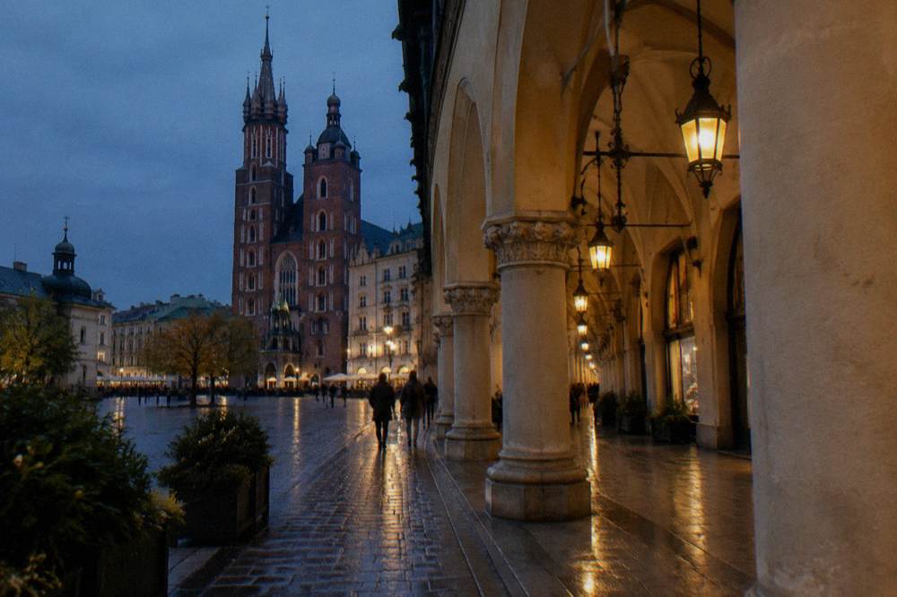 Adoquines mojados y faroles cálidos bordean las galerías del Sukiennice al anochecer, con la basílica de Santa María visible al otro lado de la plaza del mercado de Cracovia.